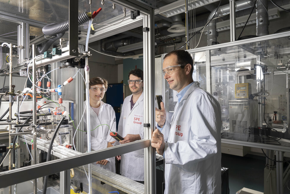 Carlijn Sebregts, Rodrigo Lira Garcia Barros en Thijs de Groot bekijken de opstelling van de alkaline elektrolyzer in het lab van de TU/e. 